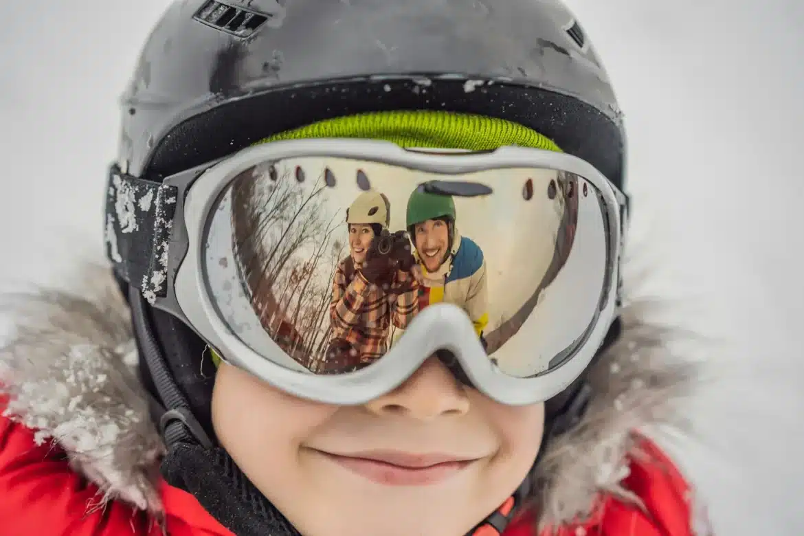A child wearing a ski helmet and goggles with a reflection of two people in the goggles smiling on the slopes