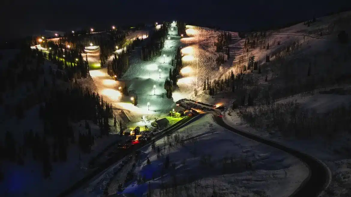 A nighttime aerial view of a ski resort with lit slopes and a parking area