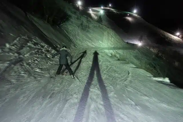 A skier on a snow covered slope at night casting a long shadow under the lights of the ski resort A skier on a snow covered slope at night casting a long shadow under the lights of the ski resort