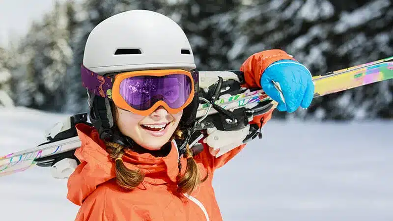 A young skier smiling while wearing a helmet goggles and holding skis on a snowy slope