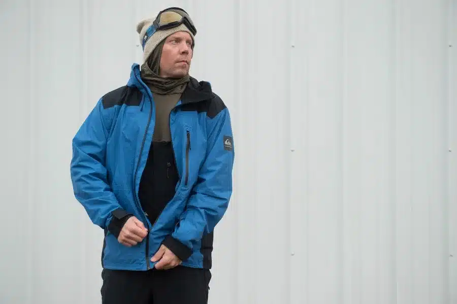 A person wearing a blue and black jacket goggles and a beige beanie standing against a white background A person wearing a blue and black jacket goggles and a beige beanie standing against a white background