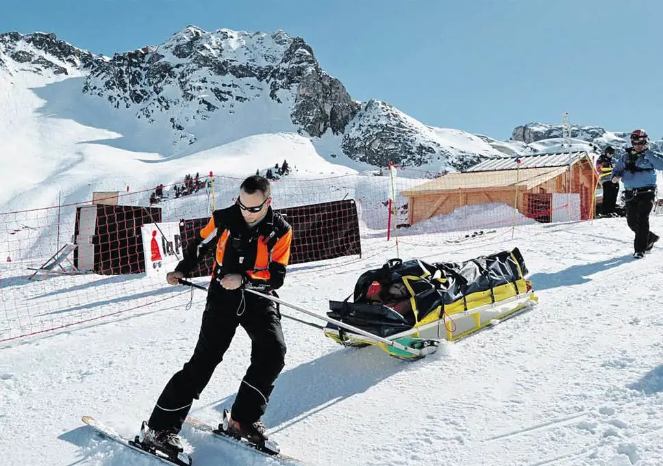 A ski patrol member pulling a rescue sled on the snow covered slopes with mountains in the background